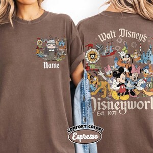 May include: Espresso-colored t-shirt featuring a front pocket design with a cartoon of a man and the word "Name". The back of the shirt displays "Walt Disney's Disneyworld Est. 1971" with Disney characters.