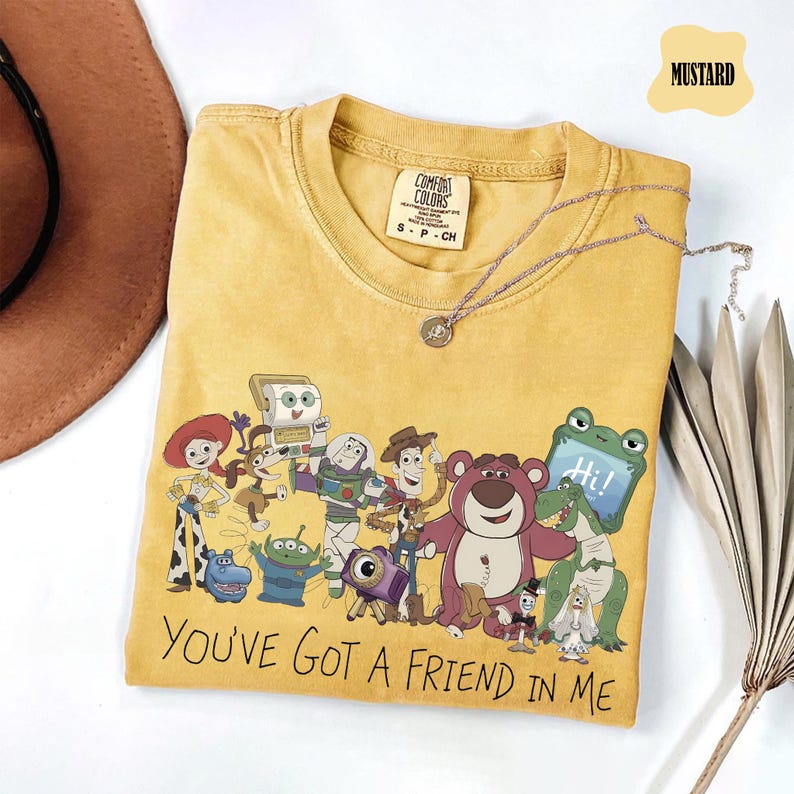 May include: A mustard yellow crew neck t-shirt featuring a graphic print of various Toy Story characters and the text "YOU'VE GOT A FRIEND IN ME". The shirt has a Comfort Colors brand label on the inside collar indicating a size small.