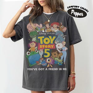 Op de afbeelding: Een antracietgrijs T-shirt met korte mouwen en een grafische print van Toy Story 5-personages, waaronder Woody, Buzz Lightyear en Jessie. De tekst luidt Disney Pixar Toy Story 5 en You've Got A Friend In Me. Een Comfort Colors Pepper-label is zichtbaar.