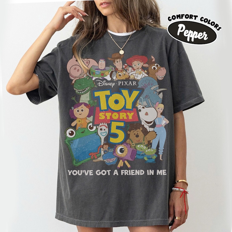 Op de afbeelding: Een antracietgrijs T-shirt met korte mouwen en een grafische print van Toy Story 5-personages, waaronder Woody, Buzz Lightyear en Jessie. De tekst luidt Disney Pixar Toy Story 5 en You've Got A Friend In Me. Een Comfort Colors Pepper-label is zichtbaar.