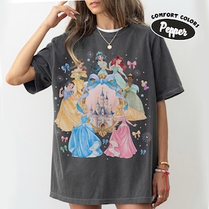 May include: Dark gray Comfort Colors t-shirt featuring a graphic of several animated princesses surrounding a castle. The princesses are in various gowns, with a large blue bow and the text "Pepper" in a black oval.