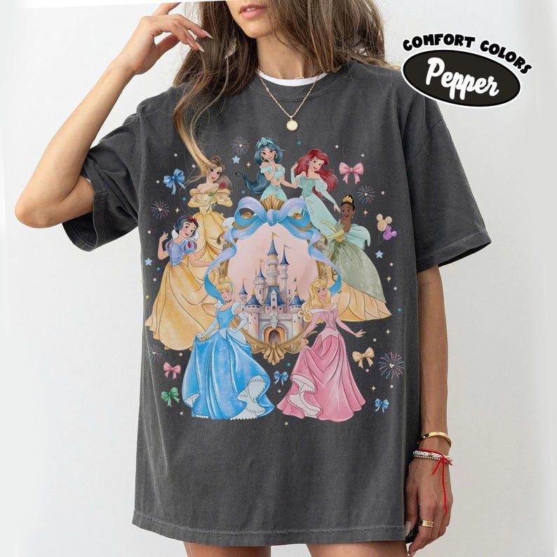 May include: Dark gray Comfort Colors t-shirt featuring a graphic of several animated princesses surrounding a castle. The princesses are in various gowns, with a large blue bow and the text "Pepper" in a black oval.