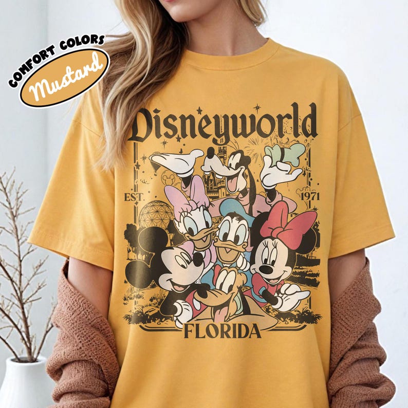 May include: Mustard yellow t-shirt featuring a graphic of Disney characters including Mickey Mouse, Minnie Mouse, Donald Duck, Daisy Duck, Goofy, and Pluto. The shirt has the text "Disneyworld Florida" and "EST. 1971".