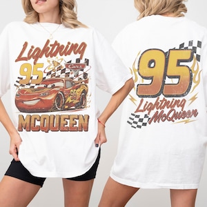 Może przedstawiać: Biała koszulka z grafiką Zygzaka McQueena z filmu Auta. Prz&oacute;d koszulki zawiera napis "Lightning 95 McQueen" i wizerunek samochodu. Tył koszulki ma duży napis "95" z napisem "Lightning McQueen" poniżej.