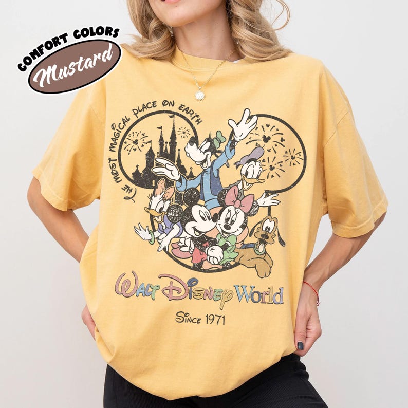 May include: Mustard-colored t-shirt featuring a graphic of Disney characters, including Mickey Mouse, Minnie Mouse, and Goofy. The design includes the text "Walt Disney World" and "Since 1971". The shirt also has the text "The Most Magical Place on Earth".