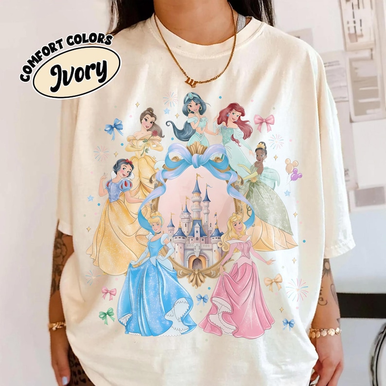 May include: Ivory-colored t-shirt featuring a colorful illustration of several princesses surrounding a castle. The design includes a blue ribbon, bows, and stars. The words "COMFORT COLORS" and "Ivory" are printed on the top left.