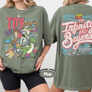 Pode incluir: Uma t-shirt verde musgo com gr&aacute;ficos de Toy Story. A frente mostra Woody, Buzz Lightyear, Rex e outras personagens com o texto "Toy Story Since 1995". As costas apresentam o log&oacute;tipo de Toy Story e o texto "To Infinity and Beyond Est. 1995".