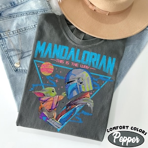 Może przedstawiać: Szara koszulka z grafiką The Mandalorian i Grogu, z napisem "MANDALORIAN" w kolorze niebieskim. Koszulka jest stylizowana z jasnoniebieskimi dżinsami i beżowym kapeluszem.