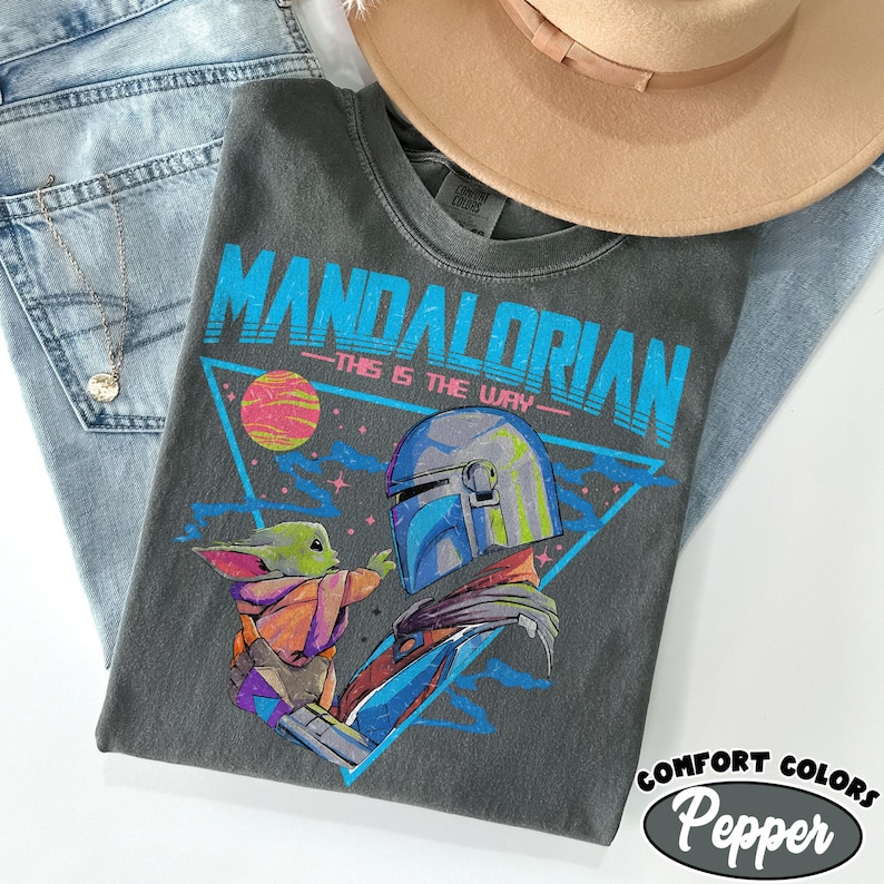 Może przedstawiać: Szara koszulka z grafiką The Mandalorian i Grogu, z napisem "MANDALORIAN" w kolorze niebieskim. Koszulka jest stylizowana z jasnoniebieskimi dżinsami i beżowym kapeluszem.