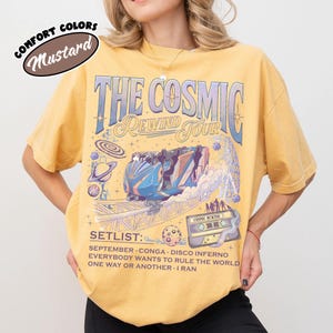 K&ouml;nnte beinhalten: Senffarbenes T-Shirt mit einem grafischen Design mit dem Text "THE COSMIC REWIND TOUR". Das Design umfasst eine Achterbahn, Planeten und die Setlist: SEPTEMBER, CONGA, DISCO INFERNO, EVERYBODY WANTS TO RULE THE WORLD, ONE WAY OR ANOTHER, I RAN.