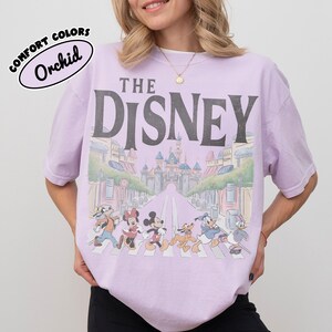 Może przedstawiać: Jasnofioletowa koszulka z szarym napisem "THE DISNEY". Koszulka przedstawia grafikę postaci Disneya przechodzących przez ulicę z zamkiem w tle. Tekst "COMFORT COLORS" i "Orchid" znajduje się w bąbelku po lewej stronie u g&oacute;ry.