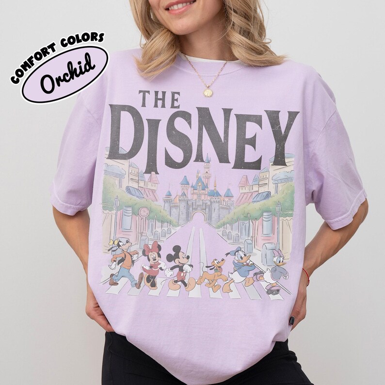 Może przedstawiać: Jasnofioletowa koszulka z szarym napisem "THE DISNEY". Koszulka przedstawia grafikę postaci Disneya przechodzących przez ulicę z zamkiem w tle. Tekst "COMFORT COLORS" i "Orchid" znajduje się w bąbelku po lewej stronie u g&oacute;ry.