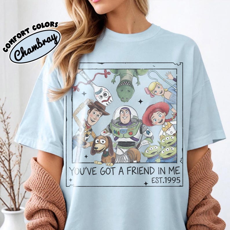 May include: Light blue t-shirt featuring a graphic of Toy Story characters within a framed design. The text "YOU'VE GOT A FRIEND IN ME EST.1995" is printed below the characters. The shirt has the text "COMFORT COLORS Chambray" in a circular design.