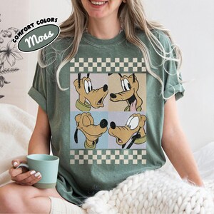 May include: A moss green t-shirt featuring a graphic of Pluto in four different poses. The design is set against a checkered background with the words "COMFORT COLORS" and "Moss" in an oval. The shirt has rolled-up sleeves.