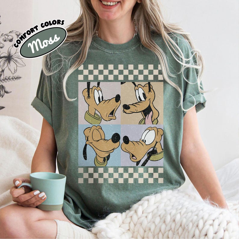 May include: A moss green t-shirt featuring a graphic of Pluto in four different poses. The design is set against a checkered background with the words "COMFORT COLORS" and "Moss" in an oval. The shirt has rolled-up sleeves.