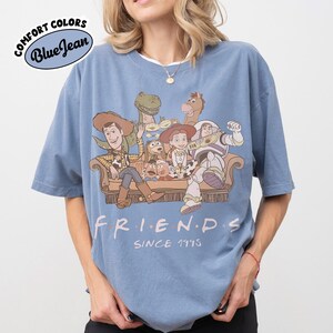 May include: Blue short-sleeved t-shirt with a graphic featuring characters from the movie Toy Story. The design includes Woody, Buzz Lightyear, and other characters sitting on a couch. The word "FRIENDS" is printed below the characters, with "SINCE 1995" underneath.