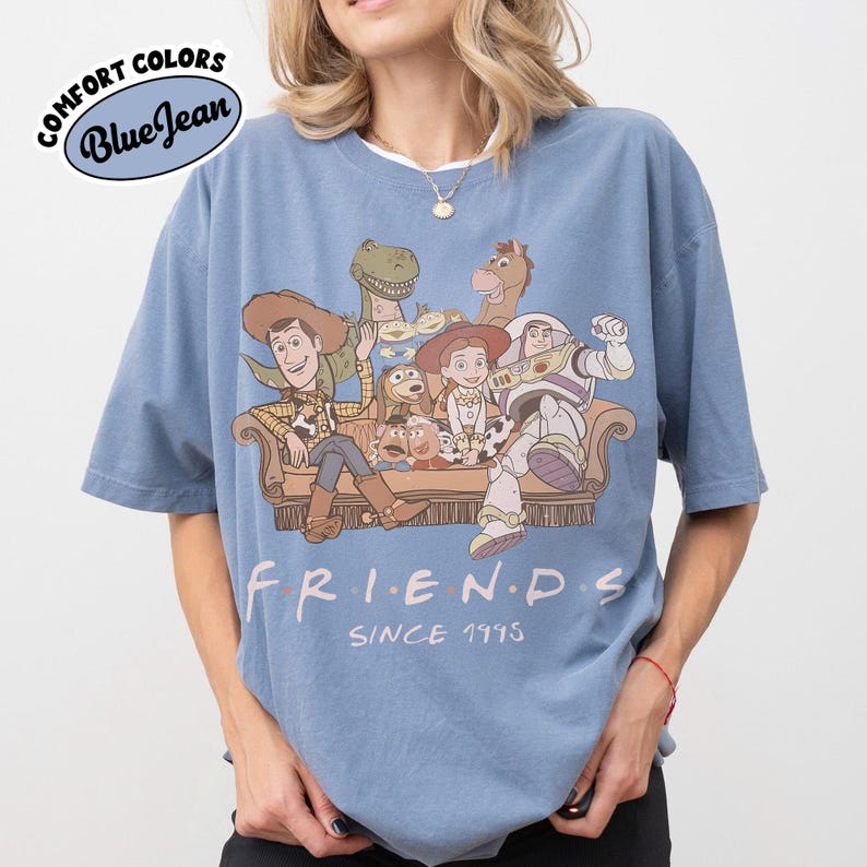 May include: Blue short-sleeved t-shirt with a graphic featuring characters from the movie Toy Story. The design includes Woody, Buzz Lightyear, and other characters sitting on a couch. The word "FRIENDS" is printed below the characters, with "SINCE 1995" underneath.