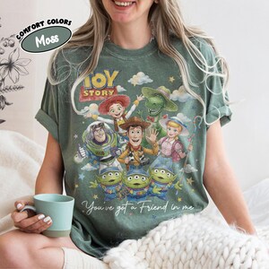 May include: Moss green t-shirt with a vibrant Toy Story character graphic, including Woody, Buzz Lightyear, and the Little Green Men. The shirt displays the text "Toy Story" and "You've got a Friend in me".