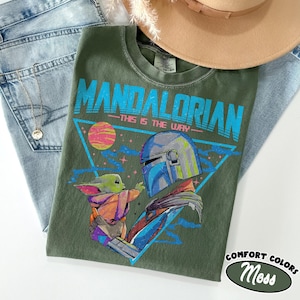 Może przedstawiać: Zielona koszulka z nadrukiem Mandalorianina i Grogu, z napisem "MANDALORIAN - THIS IS THE WAY". Koszulka jest częścią kolekcji Comfort Colors. Koszulka leży na niebieskich dżinsach i brązowym kapeluszu.