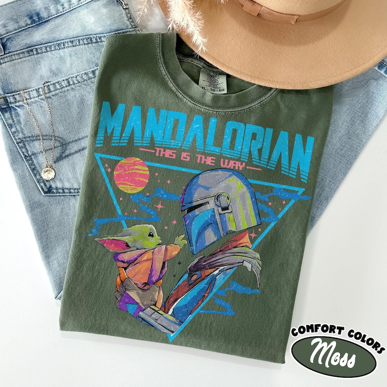 Może przedstawiać: Zielona koszulka z nadrukiem Mandalorianina i Grogu, z napisem "MANDALORIAN - THIS IS THE WAY". Koszulka jest częścią kolekcji Comfort Colors. Koszulka leży na niebieskich dżinsach i brązowym kapeluszu.