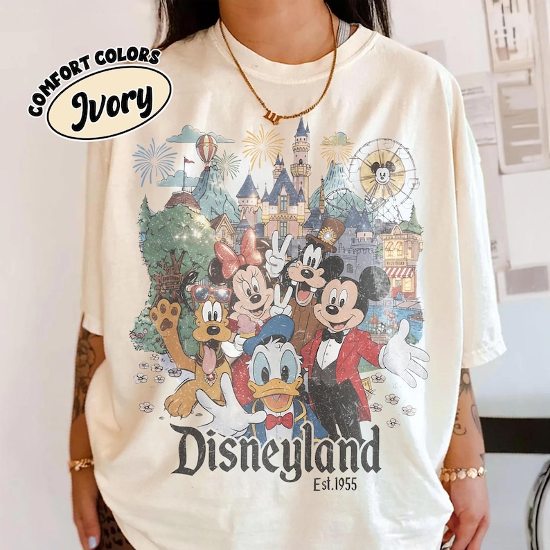 May include: Ivory-colored t-shirt featuring a colorful Disneyland graphic with Mickey Mouse, Minnie Mouse, Donald Duck, and other characters. The shirt has the words "Comfort Colors" and "Ivory" printed on it, and the word "Disneyland" is printed at the bottom.