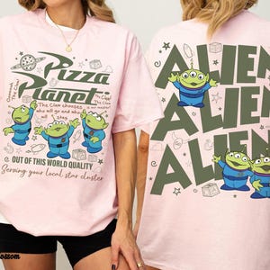 May include: Pink t-shirt featuring the Pizza Planet logo and three green aliens in blue suits. The front of the shirt has the text "Pizza Planet" and "Out of this world quality". The back of the shirt has the word "Alien" repeated.