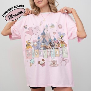 May include: A light pink t-shirt featuring a graphic print of Disneyland with a castle, Mickey Mouse, Minnie Mouse, Donald Duck, Daisy Duck, Pluto, and Goofy. The design includes cupcakes, ice cream, and the text Disneyland in a block font.