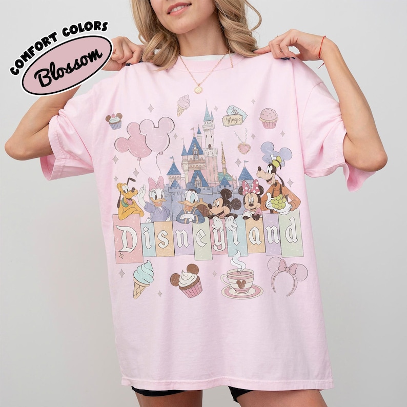 May include: A light pink t-shirt featuring a graphic print of Disneyland with a castle, Mickey Mouse, Minnie Mouse, Donald Duck, Daisy Duck, Pluto, and Goofy. The design includes cupcakes, ice cream, and the text Disneyland in a block font.