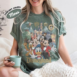 May include: Moss green t-shirt featuring a vintage Disneyland graphic with Mickey Mouse, Minnie Mouse, Donald Duck, and Goofy. The shirt has rolled-up sleeves and the words "Disneyland Est. 1955" are printed on the front.