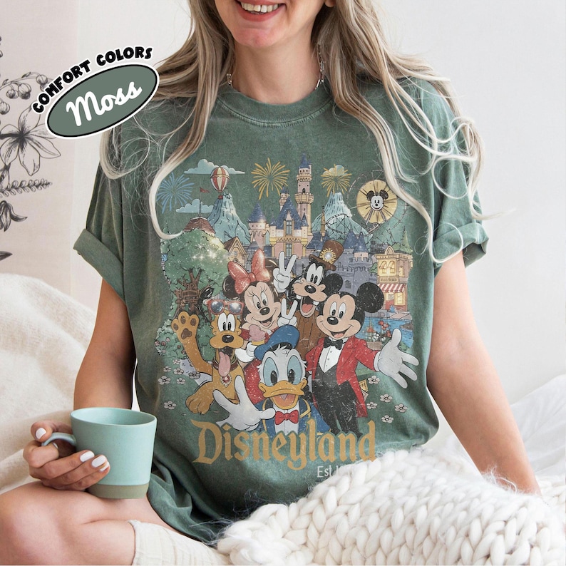 May include: Moss green t-shirt featuring a vintage Disneyland graphic with Mickey Mouse, Minnie Mouse, Donald Duck, and Goofy. The shirt has rolled-up sleeves and the words "Disneyland Est. 1955" are printed on the front.