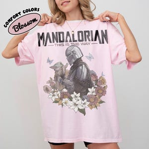May include: A light pink t-shirt featuring a graphic of The Mandalorian and Grogu surrounded by flowers and butterflies. The text "MANDALORIAN - THIS IS THE WAY" is printed across the top. The shirt also has the text "COMFORT COLORS Blossom" in a circle.