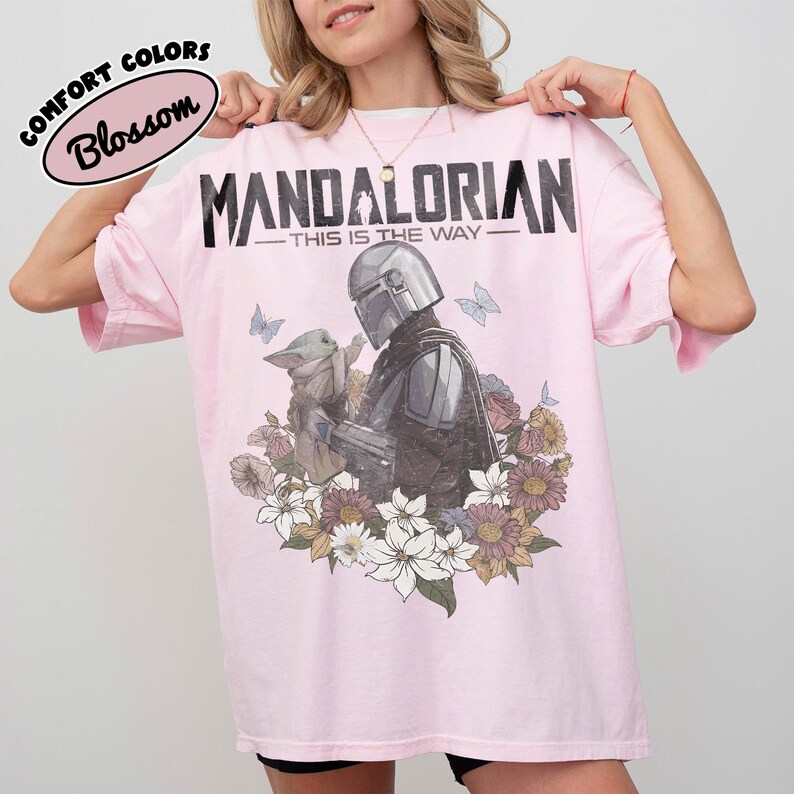 May include: A light pink t-shirt featuring a graphic of The Mandalorian and Grogu surrounded by flowers and butterflies. The text "MANDALORIAN - THIS IS THE WAY" is printed across the top. The shirt also has the text "COMFORT COLORS Blossom" in a circle.
