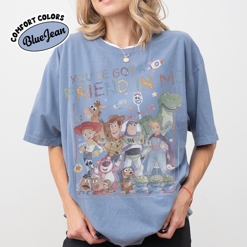 May include: A blue jean colored t-shirt featuring a graphic print of various Toy Story characters including Woody, Buzz Lightyear, Jessie, and Rex. The text "YOU'VE GOT A FRIEND IN ME" is printed above the characters in a colorful, stylized font.