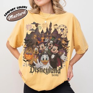 May include: A mustard yellow t-shirt featuring a graphic of Disney characters in Halloween costumes, including Mickey, Minnie, Goofy, Donald, and Pluto. The design includes a castle, fireworks, a Ferris wheel, bats, and the text Disneyland Est. 1955.