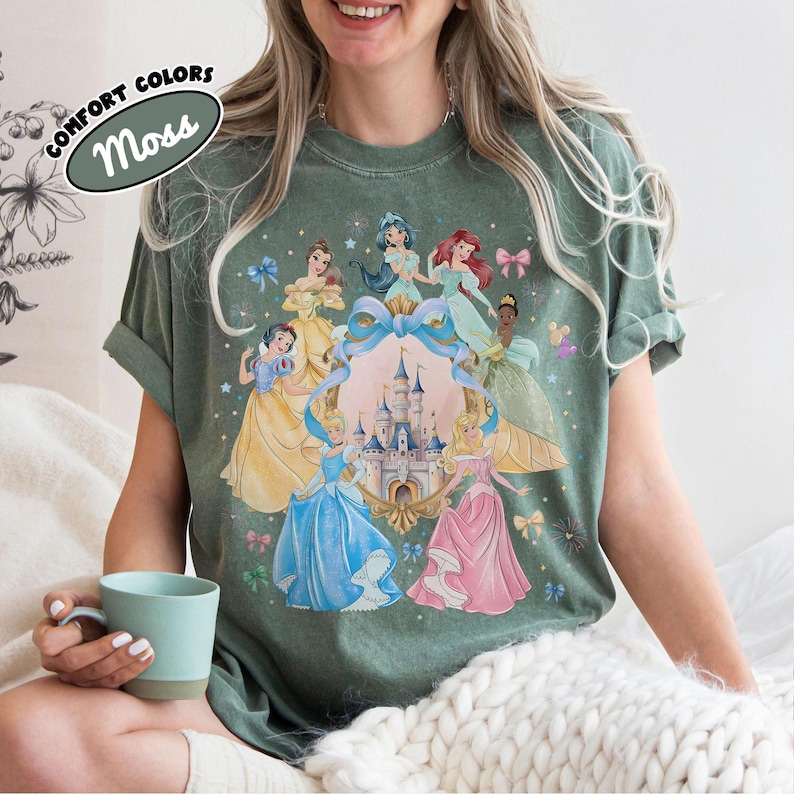 May include: Moss green t-shirt featuring a colorful graphic of several princesses surrounding a castle. The shirt has the words "Comfort Colors" and "Moss" printed on it. The shirt is a comfortable fit.