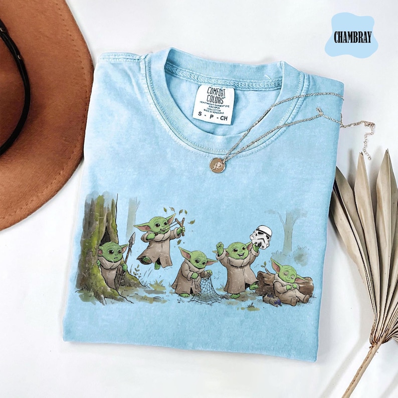 May include: A light blue t-shirt featuring a graphic print of five small green creatures in brown robes interacting with a forest environment and a white helmet. The shirt has a crew neckline and a Comfort Colors brand label at the inner collar.