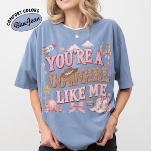 Puede incluir: Una camiseta azul con el texto YOU'RE A COWGIRL LIKE ME en letras rosas y marrones. El dise&ntilde;o incluye un cohete, un sombrero de vaquero, una herradura, una bola de discoteca, un lazo rosa y botas de vaquero, con el logo de Toy Story abajo.