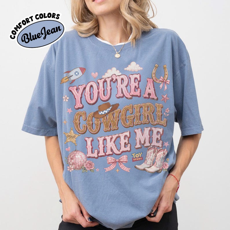 Puede incluir: Una camiseta azul con el texto YOU'RE A COWGIRL LIKE ME en letras rosas y marrones. El dise&ntilde;o incluye un cohete, un sombrero de vaquero, una herradura, una bola de discoteca, un lazo rosa y botas de vaquero, con el logo de Toy Story abajo.