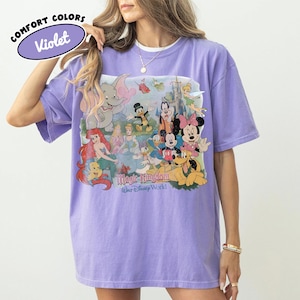 May include: Violet Comfort Colors t-shirt featuring a colorful graphic of Disney characters, including Mickey Mouse, Minnie Mouse, and Ariel. The graphic also includes the text "Magic Kingdom Walt Disney World". The shirt has short sleeves and a crew neck.