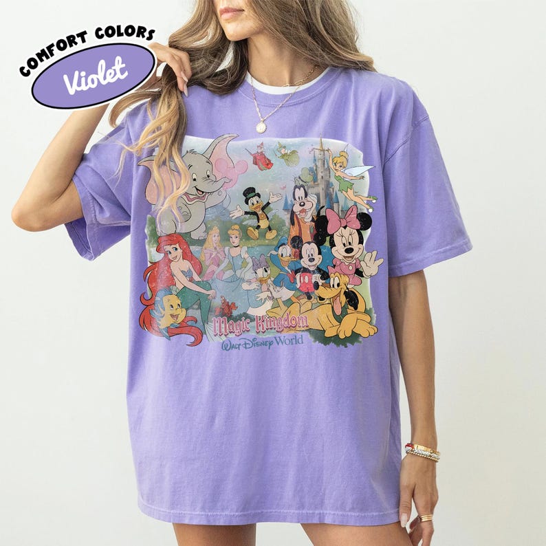 May include: Violet Comfort Colors t-shirt featuring a colorful graphic of Disney characters, including Mickey Mouse, Minnie Mouse, and Ariel. The graphic also includes the text "Magic Kingdom Walt Disney World". The shirt has short sleeves and a crew neck.