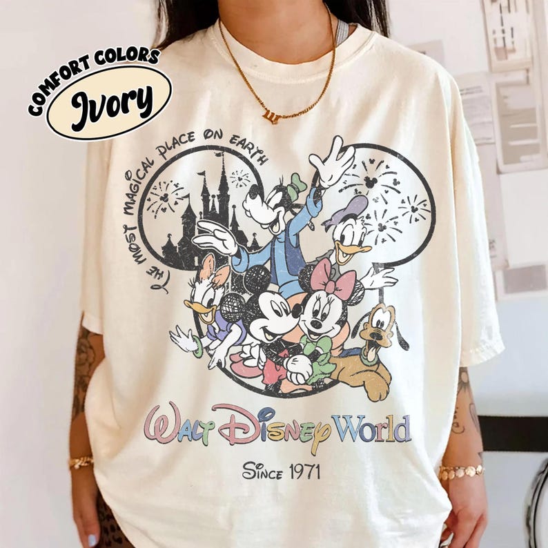 May include: Ivory-colored t-shirt featuring a graphic of Disney characters including Mickey and Minnie Mouse, Goofy, Donald and Daisy Duck, and Pluto. The shirt has the text "The Most Magical Place on Earth" and "Walt Disney World Since 1971."
