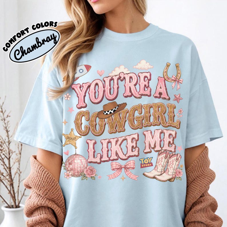 Puede incluir: Una camiseta azul claro con el texto "YOU'RE A COWGIRL LIKE ME" en rosa y marr&oacute;n. El gr&aacute;fico incluye un sombrero de vaquero, una estrella de sheriff, una bola de discoteca, una herradura, un lazo rosa y botas, con el logo "TOY STORY" abajo.