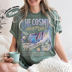 K&ouml;nnte beinhalten: Moosgr&uuml;nes T-Shirt mit einem Grafikdesign, das "THE COSMIC REWIND TOUR" und eine Achterbahn-Illustration zeigt. Die Setlist enth&auml;lt Songs wie "September" und "Everybody Wants to Rule the World".