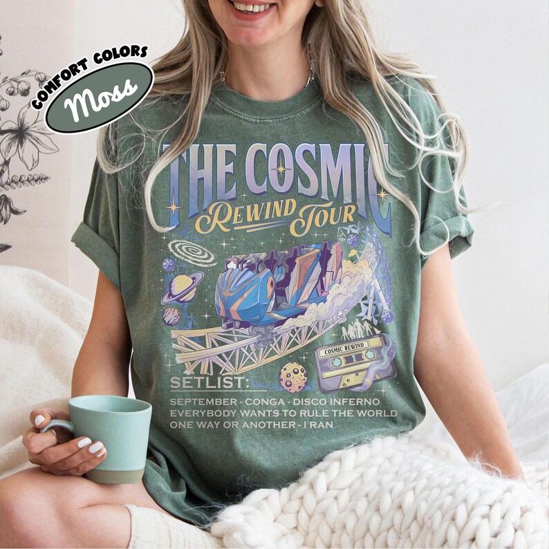 K&ouml;nnte beinhalten: Moosgr&uuml;nes T-Shirt mit einem Grafikdesign, das "THE COSMIC REWIND TOUR" und eine Achterbahn-Illustration zeigt. Die Setlist enth&auml;lt Songs wie "September" und "Everybody Wants to Rule the World".
