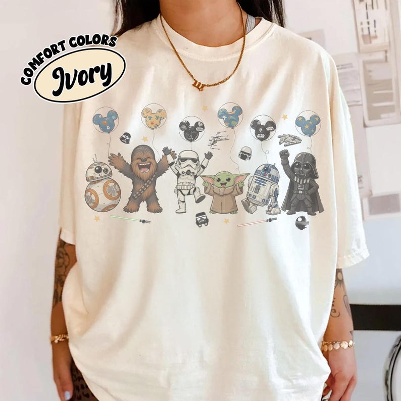 K&ouml;nnte beinhalten: Ein elfenbeinfarbenes T-Shirt mit einem Grafikdruck von Star Wars-Figuren, darunter Chewbacca, ein Stormtrooper, Grogu, R2-D2, BB-8 und Darth Vader. Die Figuren sind im Cartoon-Stil mit Luftballons und kleinen Symbolen dar&uuml;ber dargestellt.