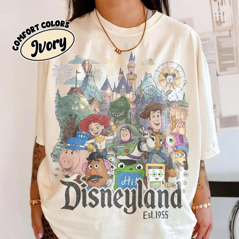May include: An ivory t-shirt featuring a graphic print of various animated characters in front of a castle and a Ferris wheel. The text "Disneyland Est. 1955" is printed in black at the bottom. A small oval label in the top left corner reads "Comfort Colors Ivory".
