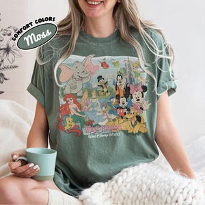 May include: Moss green t-shirt featuring a colorful graphic of Disney characters, including Dumbo, Mickey Mouse, and Ariel. The shirt has rolled-up sleeves and the words "Magic Kingdom Walt Disney World" are printed on the graphic. The shirt also has the words "Comfort Colors" and "Moss" printed on it.