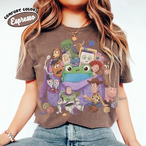 Pode incluir: Uma t-shirt de manga curta cor de caf&eacute; expresso com uma impress&atilde;o gr&aacute;fica de v&aacute;rias personagens animadas da franquia Toy Story, incluindo um sapo verde central, Buzz Lightyear, Woody e uma mochila roxa, com o texto Comfort Colors Espresso.