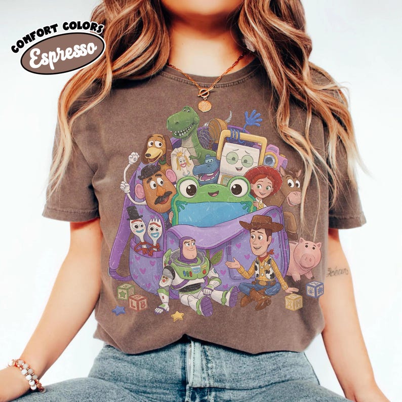 Pode incluir: Uma t-shirt de manga curta cor de caf&eacute; expresso com uma impress&atilde;o gr&aacute;fica de v&aacute;rias personagens animadas da franquia Toy Story, incluindo um sapo verde central, Buzz Lightyear, Woody e uma mochila roxa, com o texto Comfort Colors Espresso.