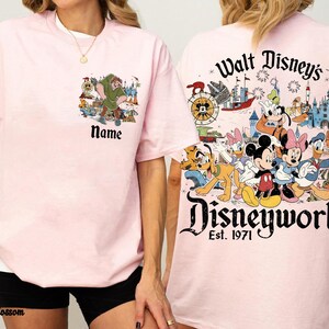 May include: Light pink t-shirts with Disney character graphics. One shirt features a cartoon scene with the text "Name", and the other displays "Walt Disney's Disneyworld Est. 1971" with various characters and a castle.